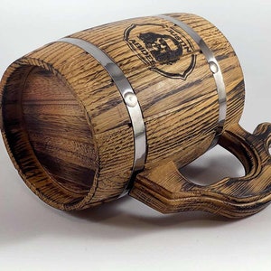 May include: A wooden beer stein with a metal band and a handle. The stein is decorated with a wood-burned design of a bearded man's face and the words "The Mustache Brigade".