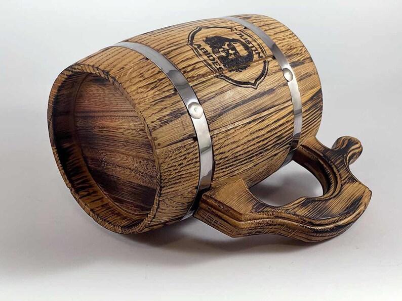May include: A wooden beer stein with a metal band and a handle. The stein is decorated with a wood-burned design of a bearded man's face and the words "The Mustache Brigade".