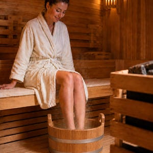 Puede incluir: Una persona con una bata blanca está sentada en una sauna, con los pies sumergidos en un baño de pies de madera. La sauna es de madera y una caja de madera es visible a la derecha. La escena está iluminada por una luz cálida y suave.
