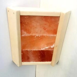 Puede incluir: Una lámpara de pared de madera de color claro con un marco rectangular. El marco contiene tres bloques rectangulares apilados de sal del Himalaya de color naranja rosado. La lámpara tiene forma trapezoidal.