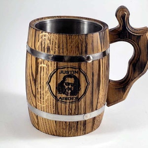 May include: A wooden beer stein with a stainless steel interior. The stein has a barrel shape with metal bands around the top and bottom. The handle is made of wood and has a decorative design. The stein is engraved with the text "Justin Abides" and a picture of a man with sunglasses.