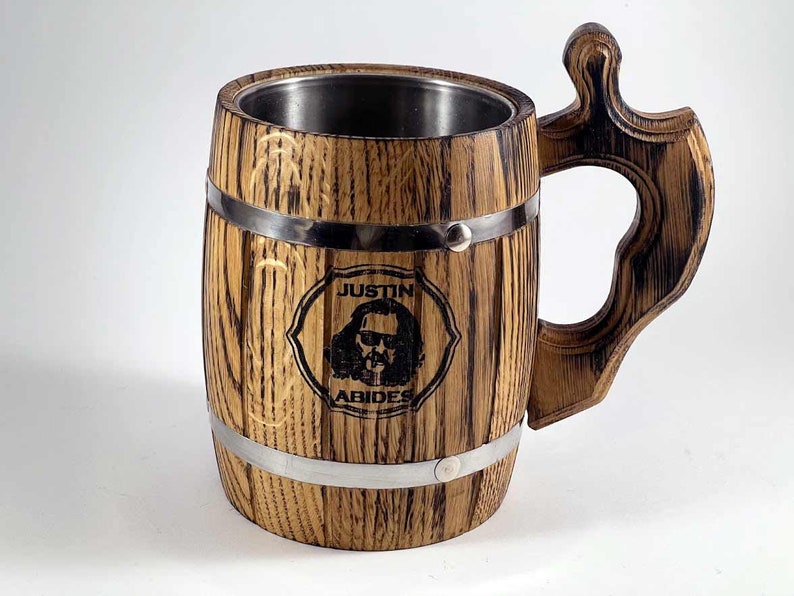 May include: A wooden beer stein with a stainless steel interior. The stein has a barrel shape with metal bands around the top and bottom. The handle is made of wood and has a decorative design. The stein is engraved with the text "Justin Abides" and a picture of a man with sunglasses.