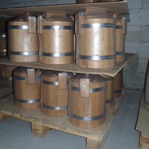 May include: A stack of wooden barrels with metal bands on a wooden pallet. The barrels are stacked two high and are all the same size. The barrels are made of light brown wood and have a natural finish.