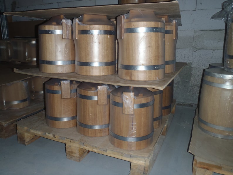 May include: A stack of wooden barrels with metal bands on a wooden pallet. The barrels are stacked two high and are all the same size. The barrels are made of light brown wood and have a natural finish.