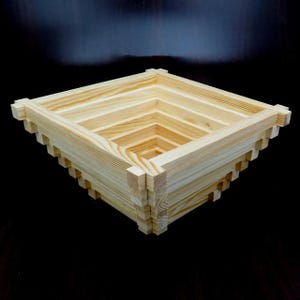 Könnte beinhalten: Eine quadratische Kiste aus Naturholz mit einem geschichteten Design. Die Kiste ist aus hellfarbenem Holz gefertigt und weist eine Reihe von verschachtelten, abnehmenden Quadraten auf. Die Kanten haben ein dekoratives, gezahntes Muster. Die Kiste ist wahrscheinlich für die Aufbewahrung oder Präsentation.