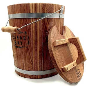 Puede incluir: Un cubo de madera con una banda de metal y una tapa de madera. El cubo tiene un asa y la tapa tiene un asa. El cubo está etiquetado con "Canoe Bay".