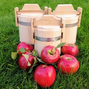 Puede incluir: Un conjunto de tres barriles de madera con bandas metálicas, rodeados de manzanas rojas sobre una superficie de hierba verde.