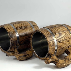 May include: Two wooden beer mugs with stainless steel interiors. The mugs are shaped like barrels and have metal bands around them. The wood is a dark brown with a wood grain pattern.