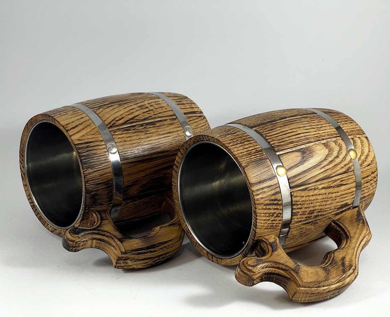 May include: Two wooden beer mugs with stainless steel interiors. The mugs are shaped like barrels and have metal bands around them. The wood is a dark brown with a wood grain pattern.