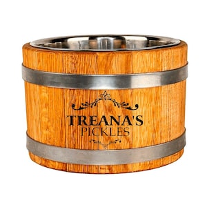 Może przedstawiać: Drewniany pojemnik w kształcie beczki z wnętrzem ze stali nierdzewnej. Pojemnik wykonany jest z jasnobrązowego drewna z dwoma srebrnymi obręczami. Z przodu nadrukowano napis "TREANA'S PICKLES".