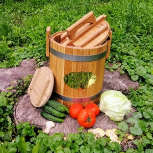May include: A wooden barrel with a lid, used for fermenting or pickling. The barrel is made of natural wood and has a metal band around the top. The lid is also made of wood and has a handle. The barrel is sitting on a wooden surface with some vegetables and a cabbage.