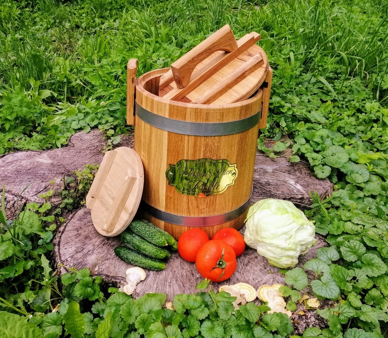 May include: A wooden barrel with a lid, used for fermenting or pickling. The barrel is made of natural wood and has a metal band around the top. The lid is also made of wood and has a handle. The barrel is sitting on a wooden surface with some vegetables and a cabbage.