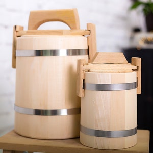 May include: Two wooden buckets with metal bands and handles. The larger bucket is in the foreground and the smaller bucket is behind it. Both buckets are made of light wood and have a natural finish.