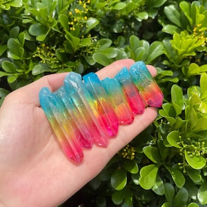 Puede incluir: Una colección de siete puntas de cristal coloridas, cada una con un degradado de colores de azul a rosa. Los cristales se sostienen en una mano, con un fondo de follaje verde.