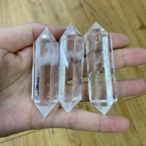 May include: Three clear quartz crystal points, also known as wands, are held in a hand. The points are smooth and polished, with a slight iridescent sheen.