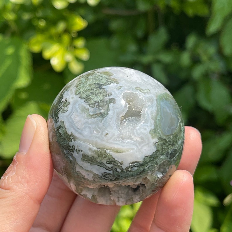 Puede incluir: Una esfera de &aacute;gata verde y blanca con un acabado natural y pulido. La esfera tiene un patr&oacute;n &uacute;nico de colores verdes y blancos que se arremolinan, con algunos peque&ntilde;os cristales transparentes visibles en el interior.