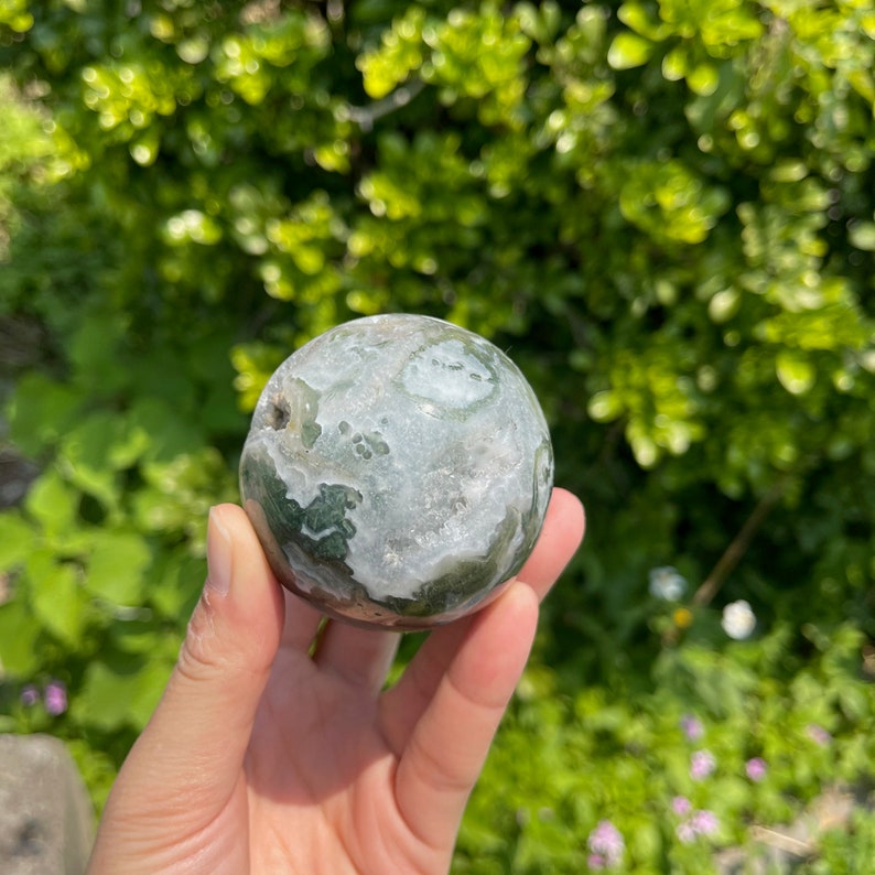 Puede incluir: Una esfera de &aacute;gata verde y blanca con un agujero perforado. La esfera es lisa y pulida, con una variedad de patrones y texturas.