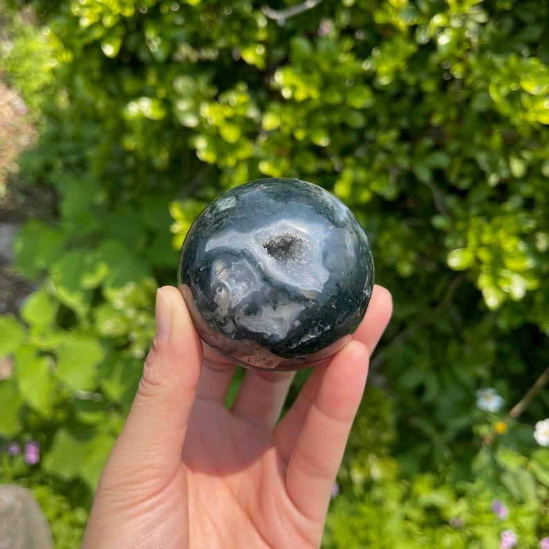 Puede incluir: Una esfera de &aacute;gata verde con un centro de drusa blanca. La esfera es lisa y pulida, y la drusa es brillante. La esfera tiene un di&aacute;metro de aproximadamente 5 cm.