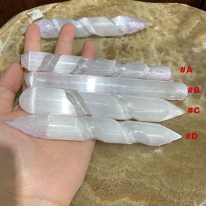 May include: Four white selenite wands with a twisted design are shown. Each wand tapers to a point at one end. The wands are held in a hand, and the background is a light brown surface. The wands are labelled with the letters A, B, C, and D.