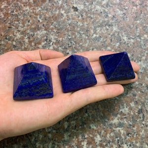 Peut inclure: Trois pyramides en lapis-lazuli, une carrée et deux triangulaires, sur une main. Les pyramides sont d'un bleu profond avec des stries blanches.