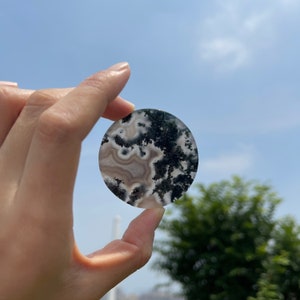 Könnte beinhalten: Ein runder, polierter Stein mit einem einzigartigen Muster aus weißen, grauen und dunkelgrünen Einschlüssen. Der Stein wird vor einem blauen Himmel gehalten, um seine natürliche Schönheit und sein komplexes Design zu zeigen. Der Durchmesser des Steins beträgt ca. 7,6 cm.