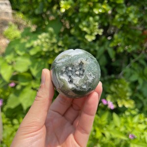 Puede incluir: Una esfera de &aacute;gata verde y blanca con una druza de cristal natural en su interior. La esfera es lisa y pulida, y la druza es brillante y blanca.