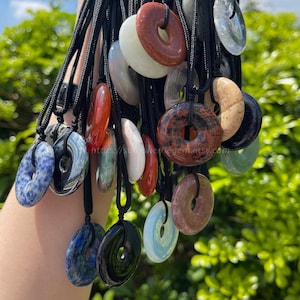 Könnte beinhalten: Eine Sammlung von Donut-Anhängern aus Edelsteinen an schwarzen Kordeln. Die Anhänger sind in verschiedenen Farben erhältlich, darunter Rot, Blau, Weiß und Schwarz. Die Anhänger haben ein Loch in der Mitte. Der Hintergrund ist verschwommenes grünes Laub.