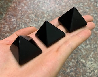 May include: Three black obsidian pyramids, each with a smooth, polished surface. The pyramids are arranged in a row on a hand, with the largest pyramid at the back and the smallest pyramid at the front.