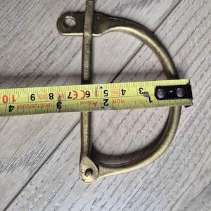 Könnte beinhalten: Ein D-Ring-Schäkel aus Messing, ca. 7,6 cm lang, mit Stift und Öse. Ein gelbes Maßband liegt über dem Schäkel und zeigt das Maß in Zentimetern an. Der Schäkel liegt auf einer Holzoberfläche.