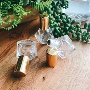 May include: Three clear glass roller bottles with gold lids on a wooden surface. The bottles are faceted and have a diamond-like shape.