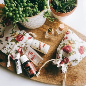 May include: A wooden cutting board displays essential oil products, including small bottles and a lip balm, alongside two floral drawstring bags. Two potted plants, a string of pearls and an aloe vera plant, are also visible.