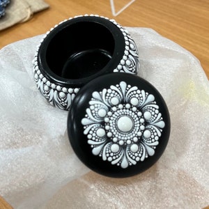 Puede incluir: Caja de baratijas redonda negra con tapa, decorada con patrones de puntos blancos. La tapa presenta un diseño floral central. La caja está abierta, revelando un interior negro. La caja descansa sobre una superficie blanca texturizada.
