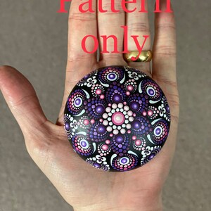 May include: A purple and pink mandala pattern painted on a round stone. The stone is held in a hand.