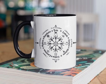 May include: White ceramic mug with a black handle and a black rim. The mug features a black and white illustration of a wheel with eight spokes and eight points. The points are labeled with the names of eight pagan holidays: Yule, Imbolc, Ostara, Beltane, Lithia, Lammas, Mabon, and Samhain. The dates of each holiday are also listed.