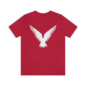 May include: Red short-sleeved t-shirt with a white stylized image of a bird with its wings spread.