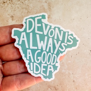 Puede incluir: Una pegatina con la forma de Devon, con el texto "Devon is Always a Good Idea" en letras blancas sobre un fondo turquesa. La pegatina está sujeta en una mano.