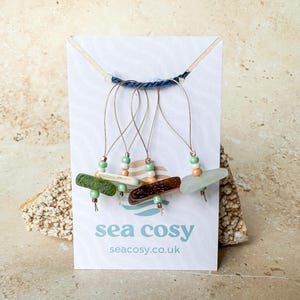 Könnte beinhalten: Ohrringe aus Meeresglas mit silberfarbenen Drähten und bunten Perlen. Sie werden auf einer weißen Karte mit dem Text "sea cosy" und einer Website-Adresse präsentiert. Die Meeresglasteile sind in verschiedenen Farben gehalten.