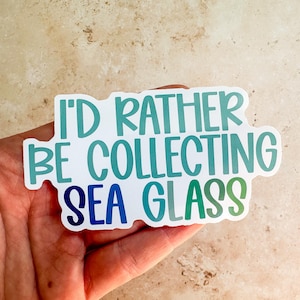 May include: A white sticker with the text "I'D RATHER BE COLLECTING SEA GLASS" in teal and blue lettering. The sticker is held in a hand against a neutral background.