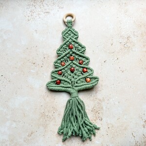 Könnte beinhalten: Ein grüner Makramee-Weihnachtsbaum-Wandbehang mit einem Holzring oben und braunen Holzperlen als Ornamenten.