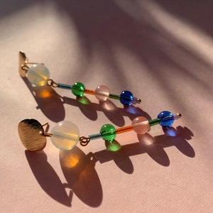 Könnte beinhalten: Ein Paar goldene Ohrringe mit einem langen, baumelnden Design. Jeder Ohrring hat oben eine runde, goldene Scheibe, gefolgt von einer weißen Opalperle und einer Reihe von bunten Glasperlen in Grün-, Blau- und Rosatönen.