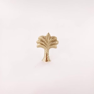 May include: A gold-colored, palm tree-shaped cabinet knob. The knob features a detailed design of palm fronds and a textured trunk. The hardware is likely made of metal and is suitable for furniture or decorative use.