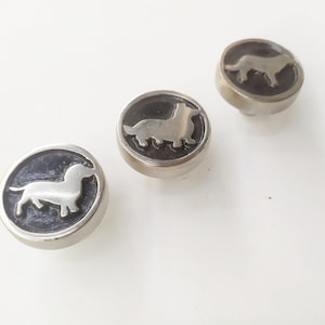 Puede incluir: Tres botones plateados con fondo negro y una silueta de dachshund blanca.