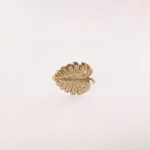 Puede incluir: Un broche dorado en forma de hoja. El broche es una representación detallada de una hoja de monstera, con intrincados patrones de venas y secciones recortadas. El broche está sobre un fondo blanco.