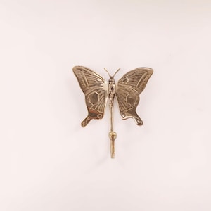 Puede incluir: Un colgador de pared con forma de mariposa de color latón. La mariposa tiene patrones de alas detallados y un pequeño gancho que se extiende desde su cuerpo. El gancho está diseñado para colgar objetos. Las alas de la mariposa están extendidas.