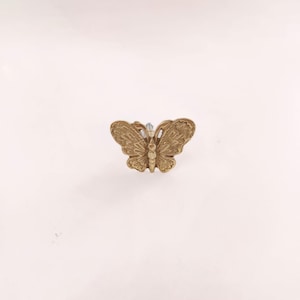 May include: A detailed, golden-colored butterfly-shaped cabinet knob. The butterfly has intricate wing patterns and is mounted on a silver-colored post. The knob is designed for furniture and adds a decorative touch.