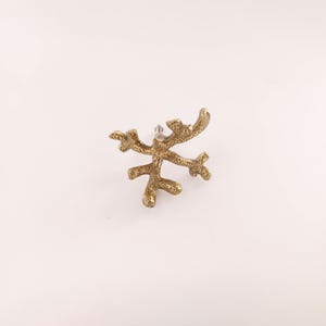 May include: A gold-coloured, coral-shaped earring stud. The earring has a textured surface and a post for insertion. The design resembles a small branch or antler, with multiple points extending outwards. The earring is set against a white background.