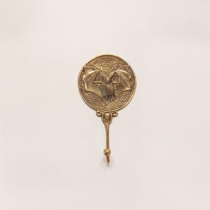 Pode incluir: Um gancho de parede de metal dourado com uma placa redonda decorativa que apresenta um design em relevo de um morcego com as asas abertas.