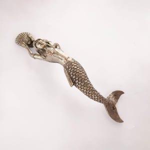May include: A silver-colored mermaid figurine, holding a seashell. The mermaid has detailed scales and flowing hair. The tail curves gracefully, and the figurine is set against a white background.