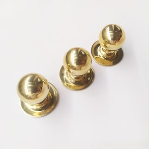 May include: Three gold-toned round cabinet knobs with a smooth, shiny finish. Each knob has a round base and a raised, spherical top.