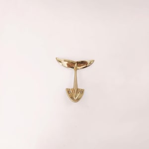 May include: A brass whale tail wall hook. The hook has a detailed whale tail design with a central stem and a mounting plate with two screw holes. The brass has a polished finish, reflecting light.